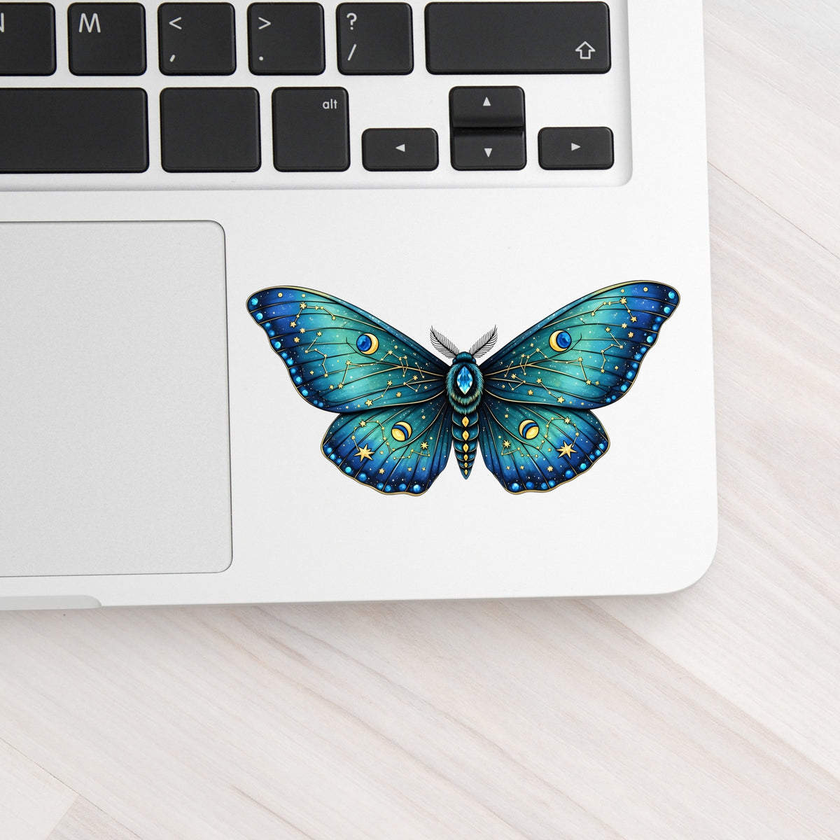 Teal Celestial Moth UV DTF sticker applied to a silver laptop beside the keyboard