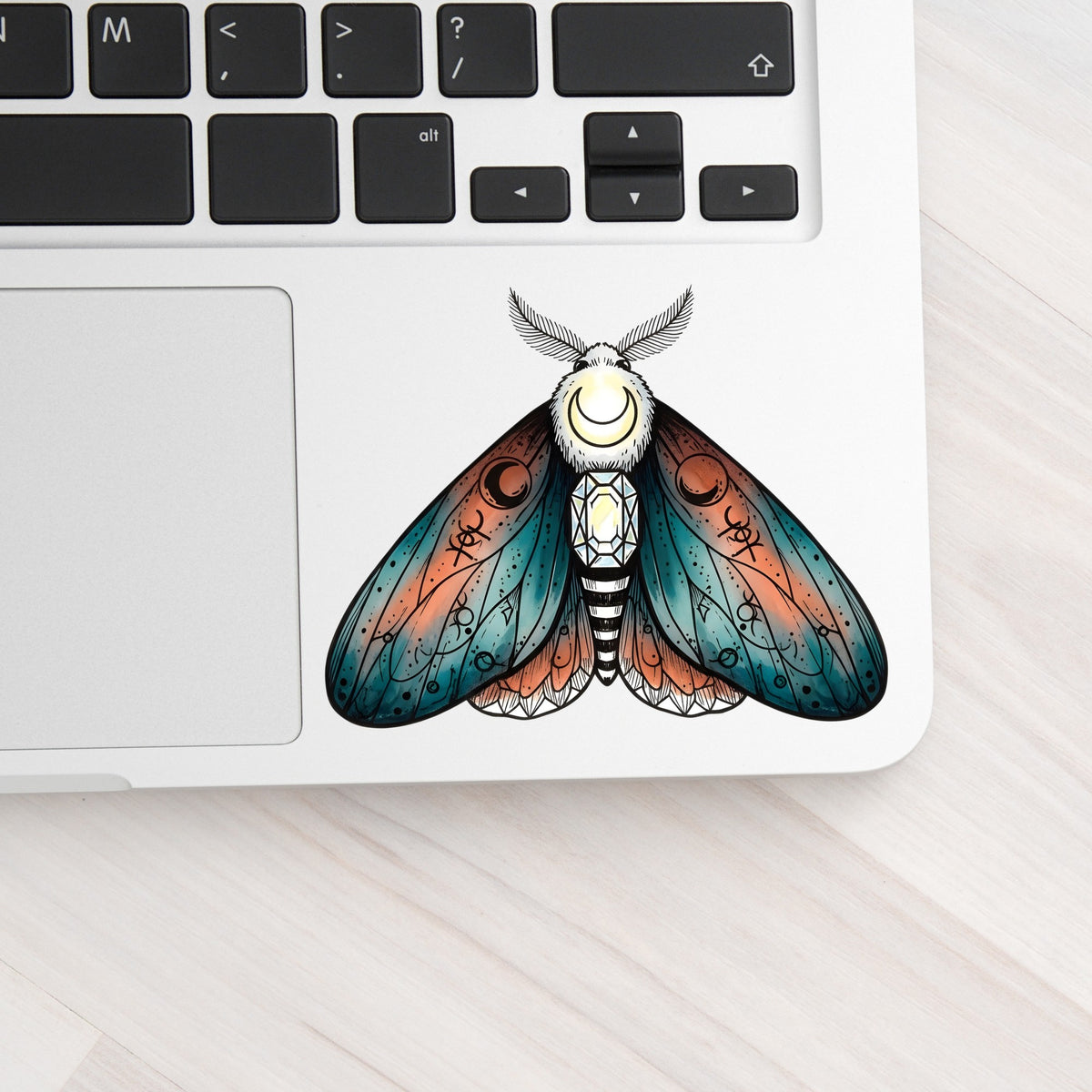 Coral Teal Crescent Moon Moth UV DTF sticker applied to a silver laptop beside the keyboard