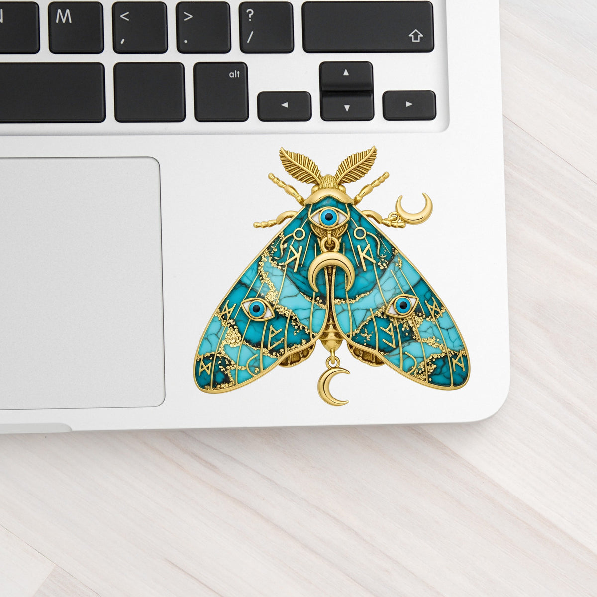 Teal Evil Eye Moth UV DTF sticker applied to a silver laptop beside the keyboard