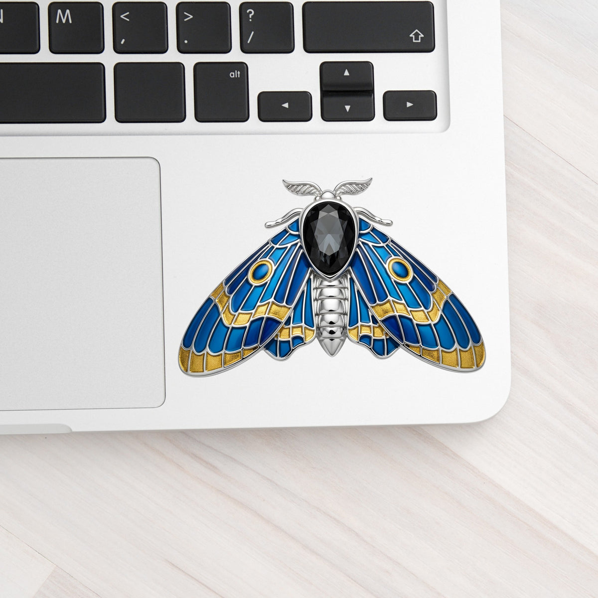 Silver Blue Enamel Moth UV DTF sticker applied to a silver laptop beside the keyboard