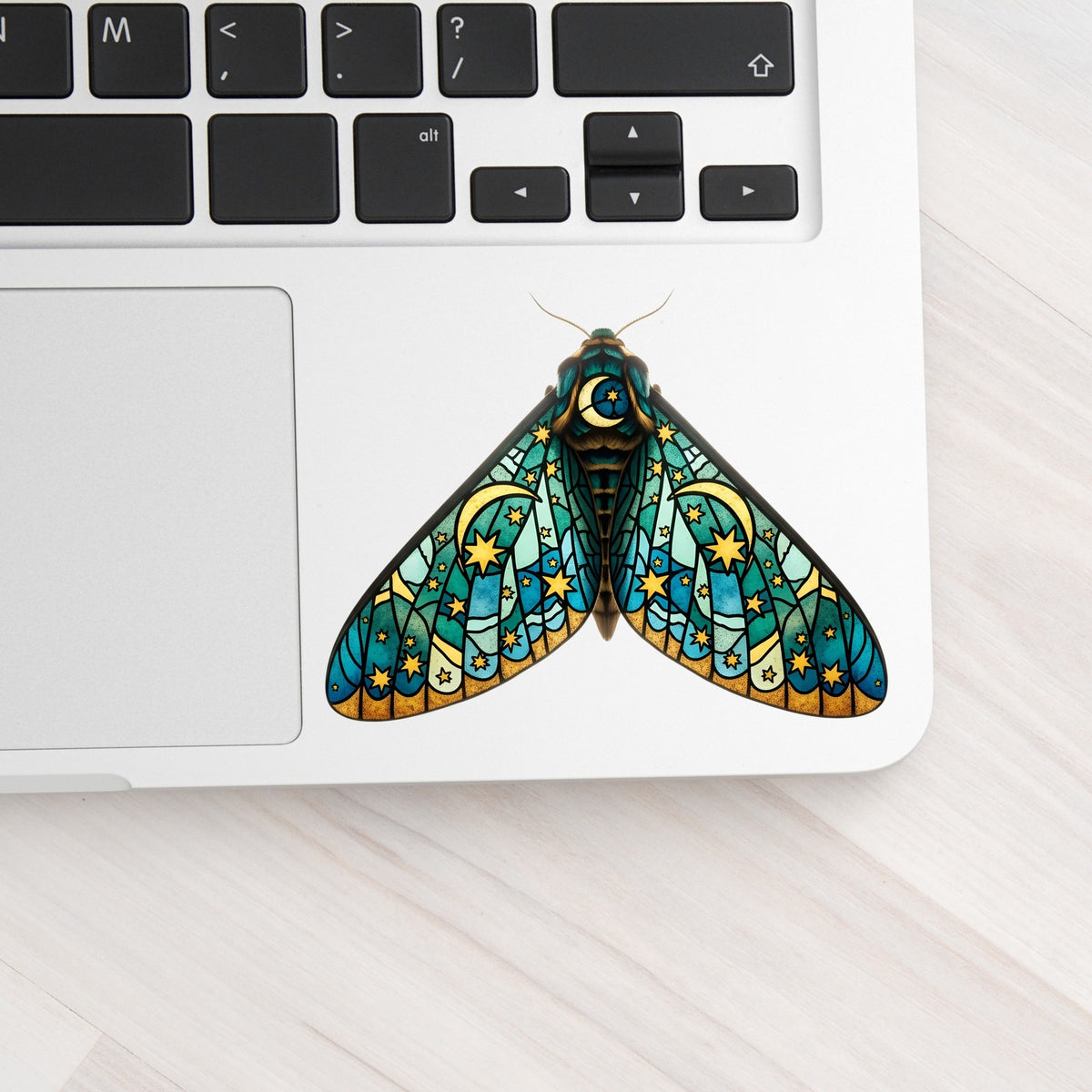 Stained Glass Moon Moth UV DTF sticker applied to a silver laptop beside the keyboard