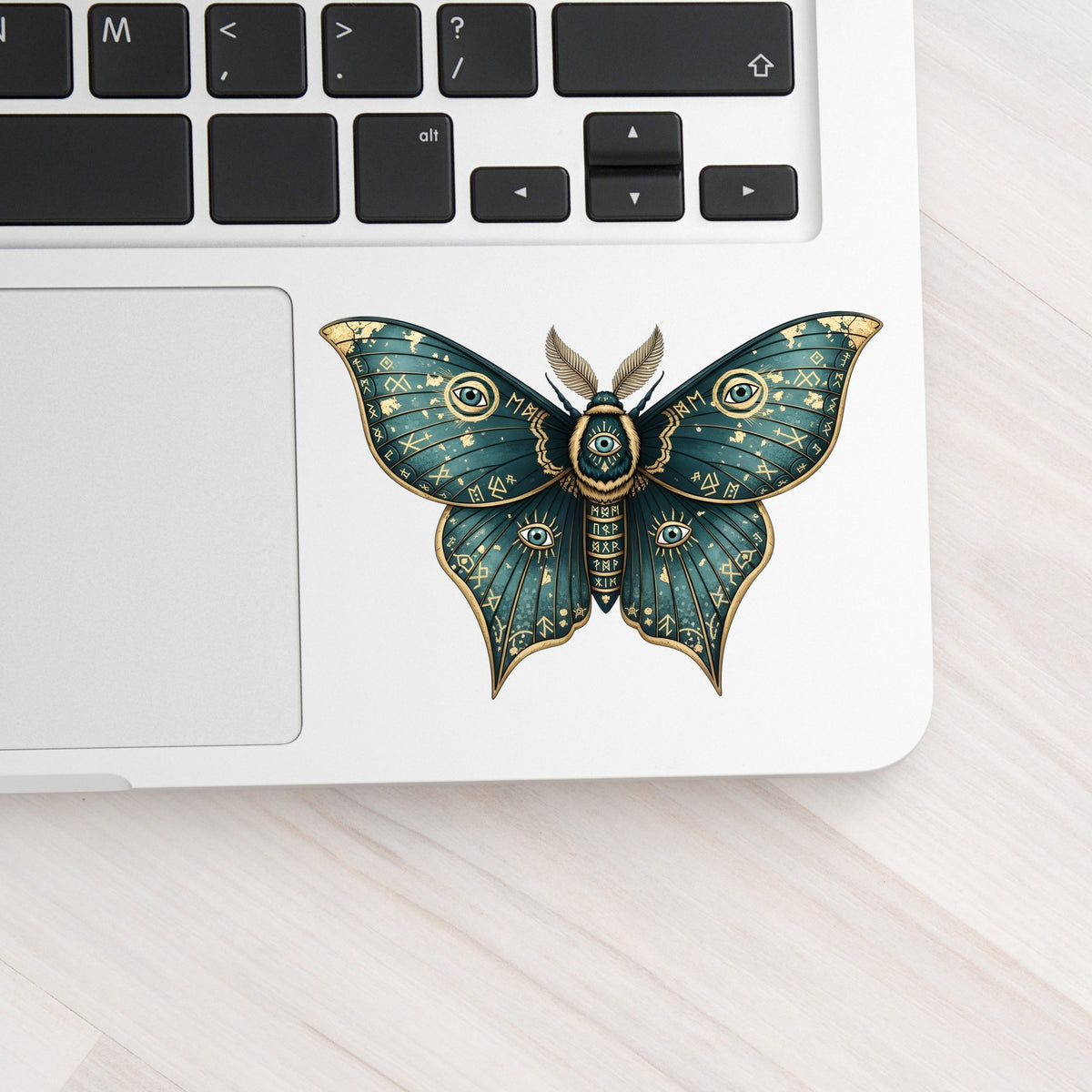 Teal Rune Moth All Seeing Eye UV DTF sticker applied to a silver laptop beside the keyboard