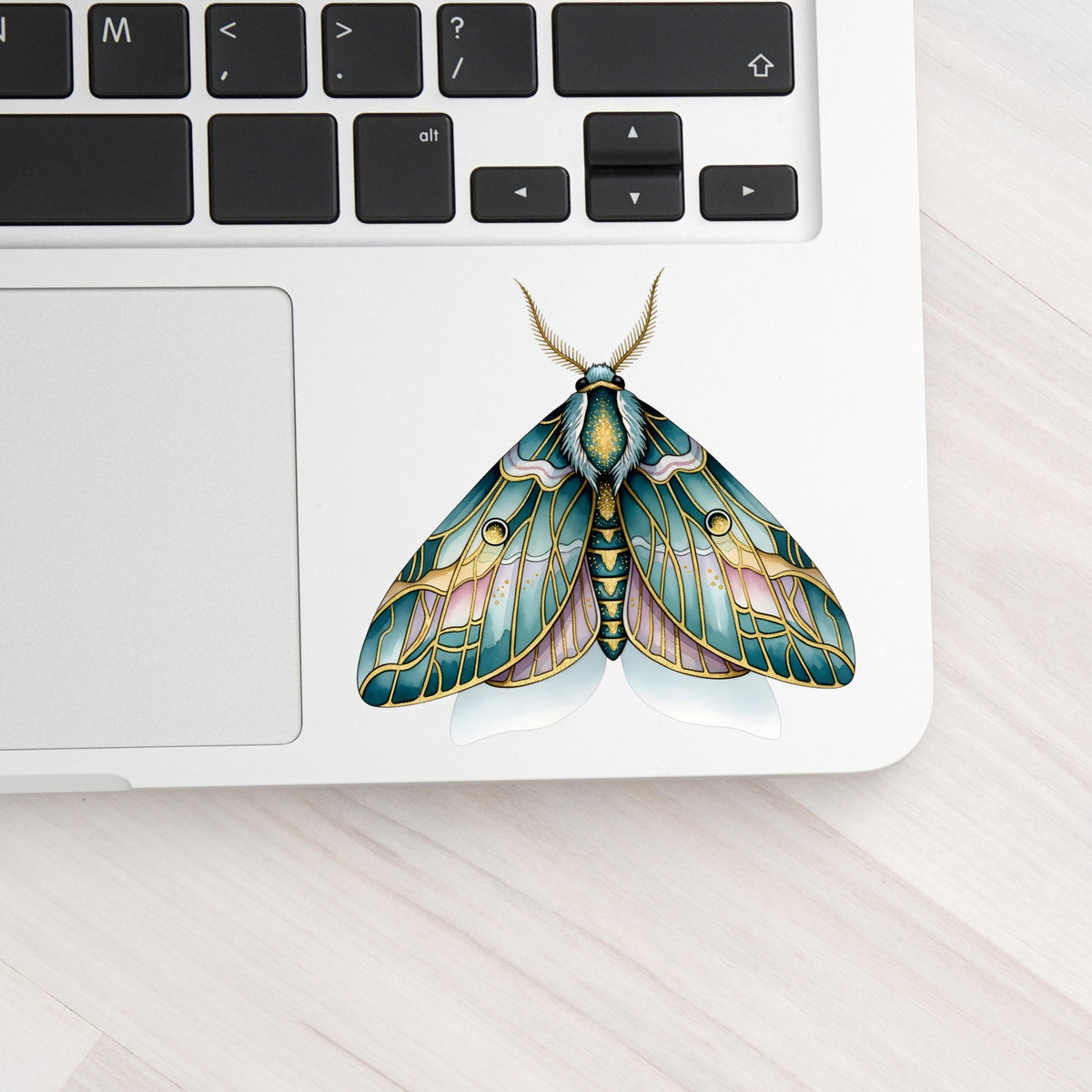 Pastel Teal Pink Moth UV DTF sticker applied to a silver laptop beside the keyboard
