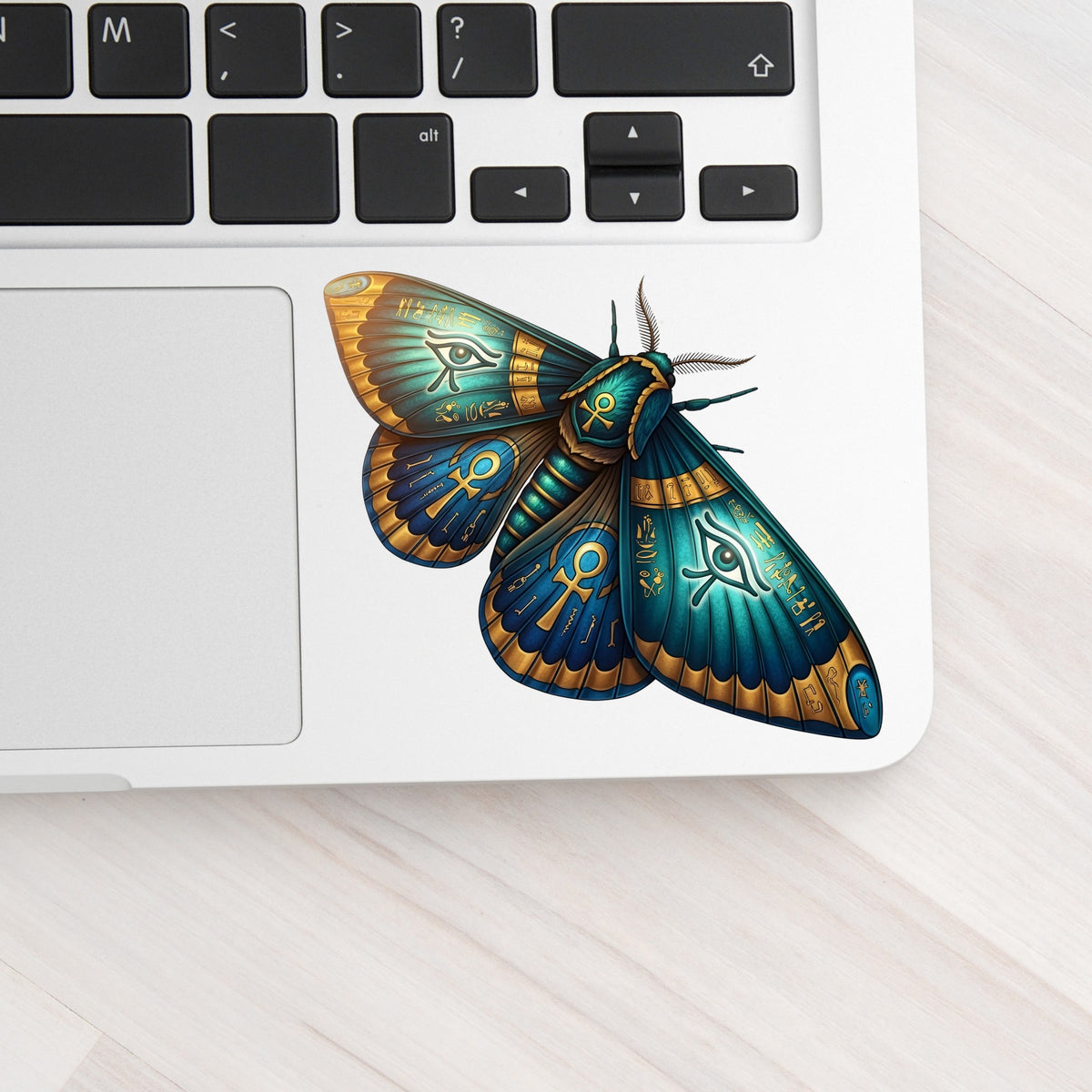 Egyptian Eye Of Horus Moth UV DTF sticker applied to a silver laptop beside the keyboard