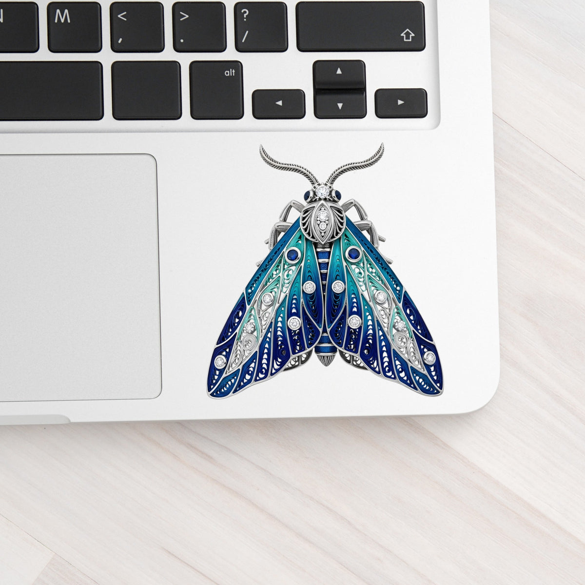Silver Filigree Blue Moth UV DTF sticker applied to a silver laptop beside the keyboard