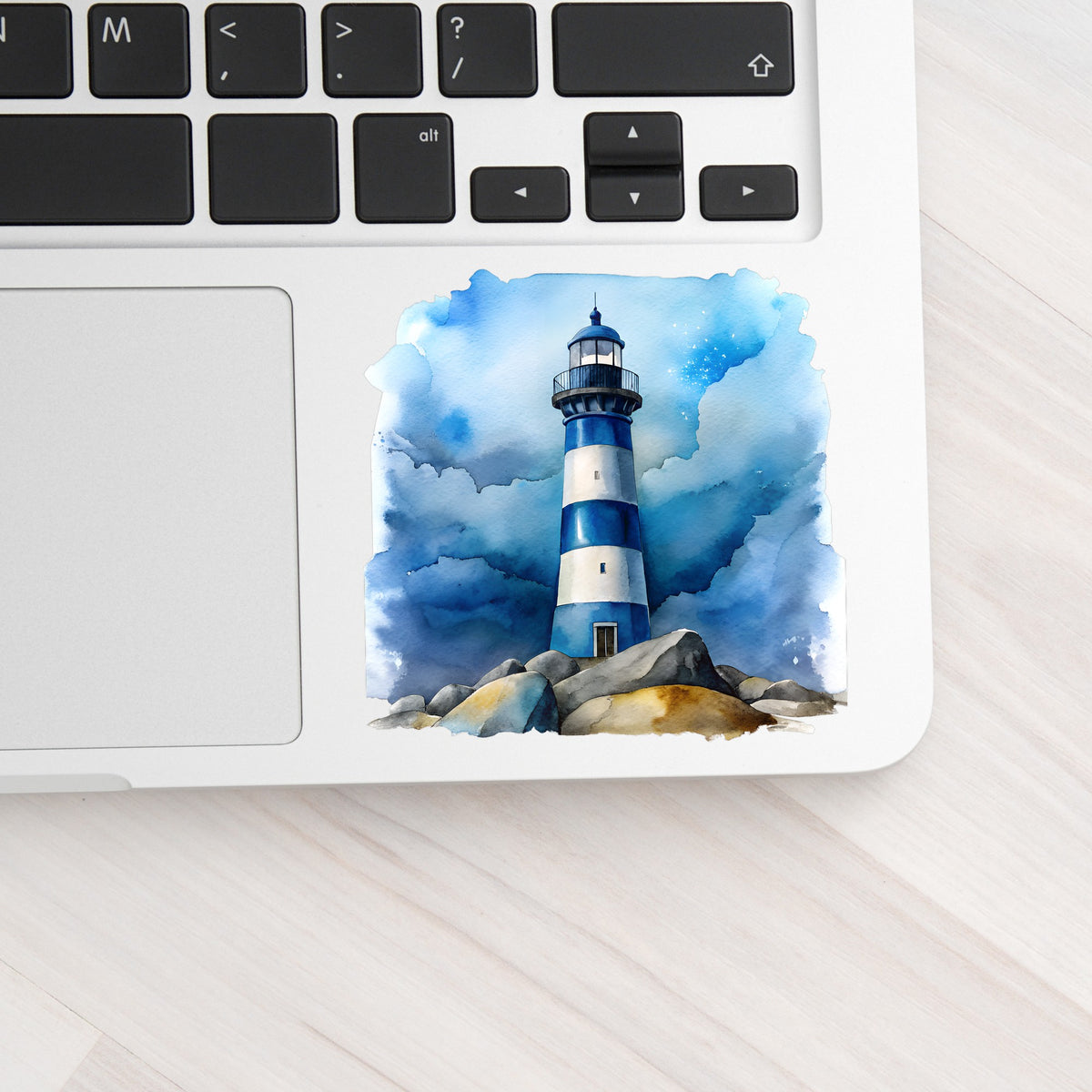 Blue Striped Lighthouse On Rocks UV DTF sticker applied to a silver laptop beside the keyboard