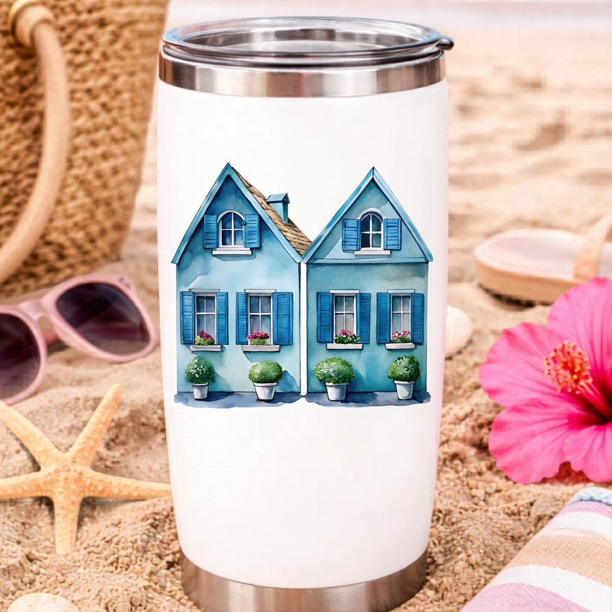 Two Blue Coastal Cottages UV DTF sticker applied to a white travel tumbler on beach sand