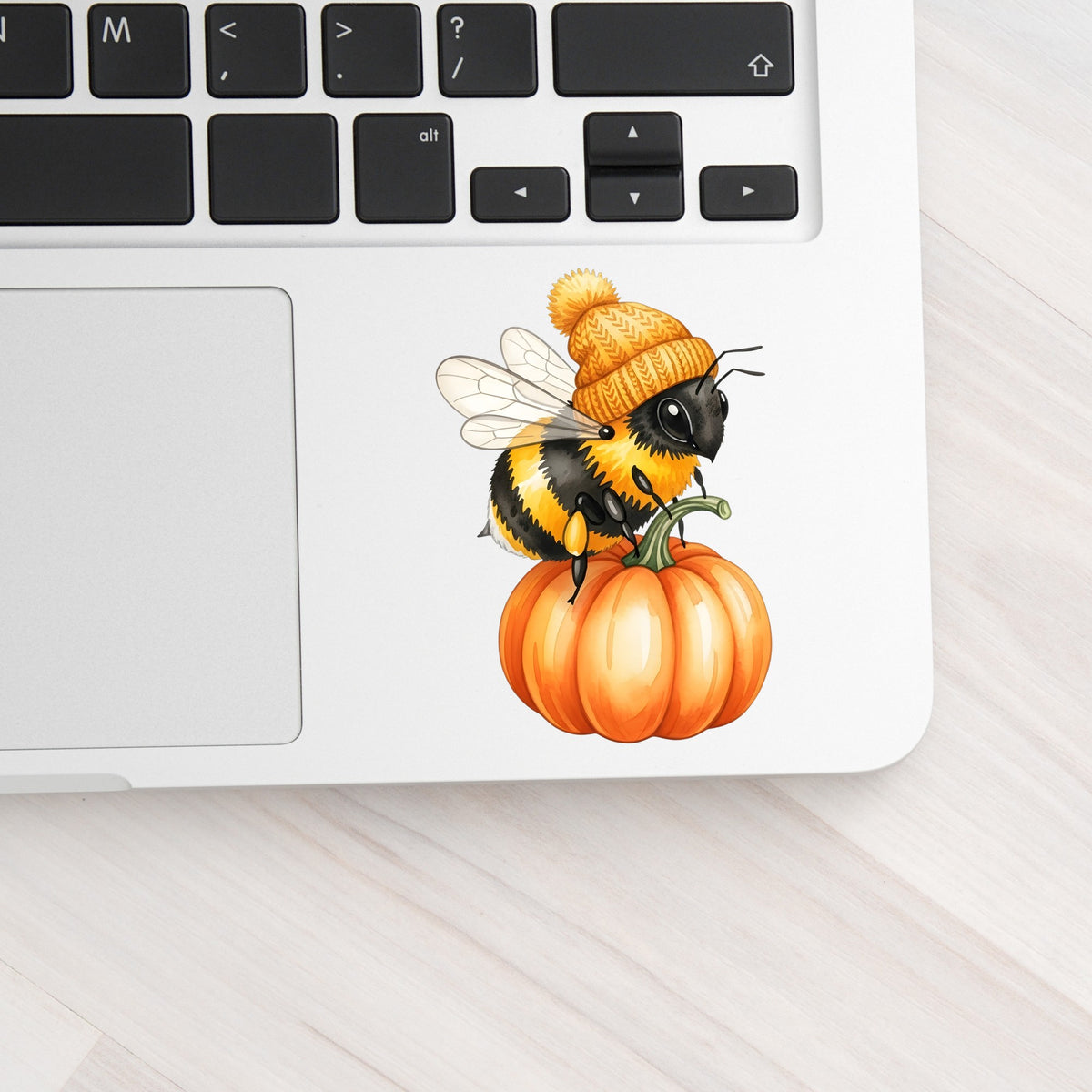 Bee In Orange Beanie On Pumpkin UV DTF sticker applied to a silver laptop beside the keyboard