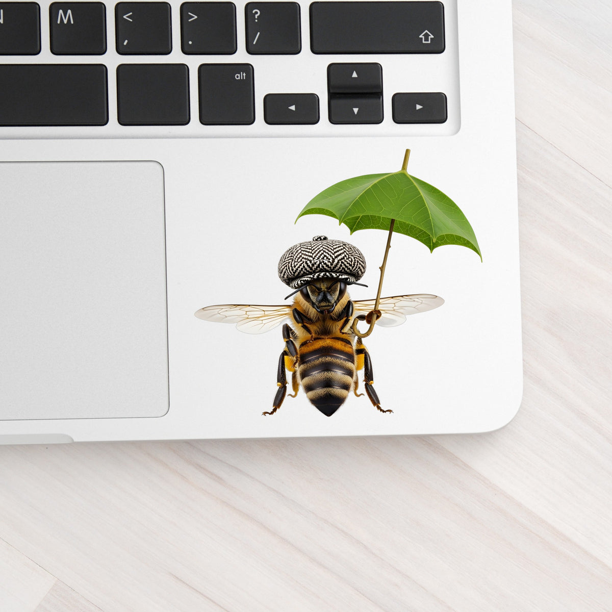 Bee In Flat Cap With Leaf Umbrella UV DTF sticker applied to a silver laptop beside the keyboard