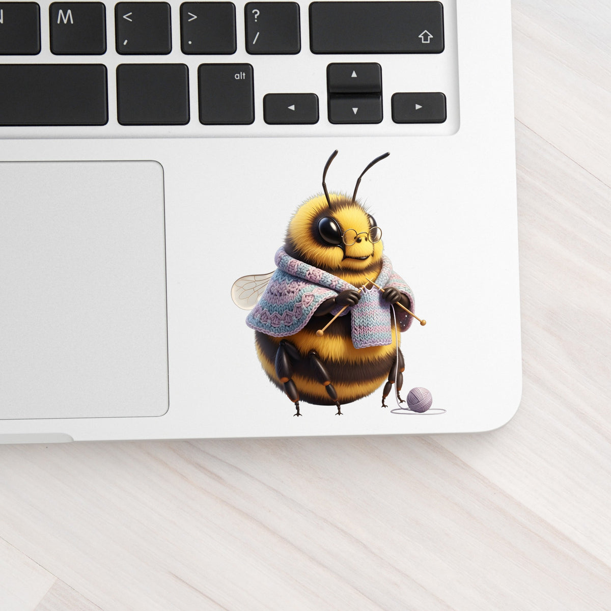 Bee Knitting In Shawl UV DTF sticker applied to a silver laptop beside the keyboard