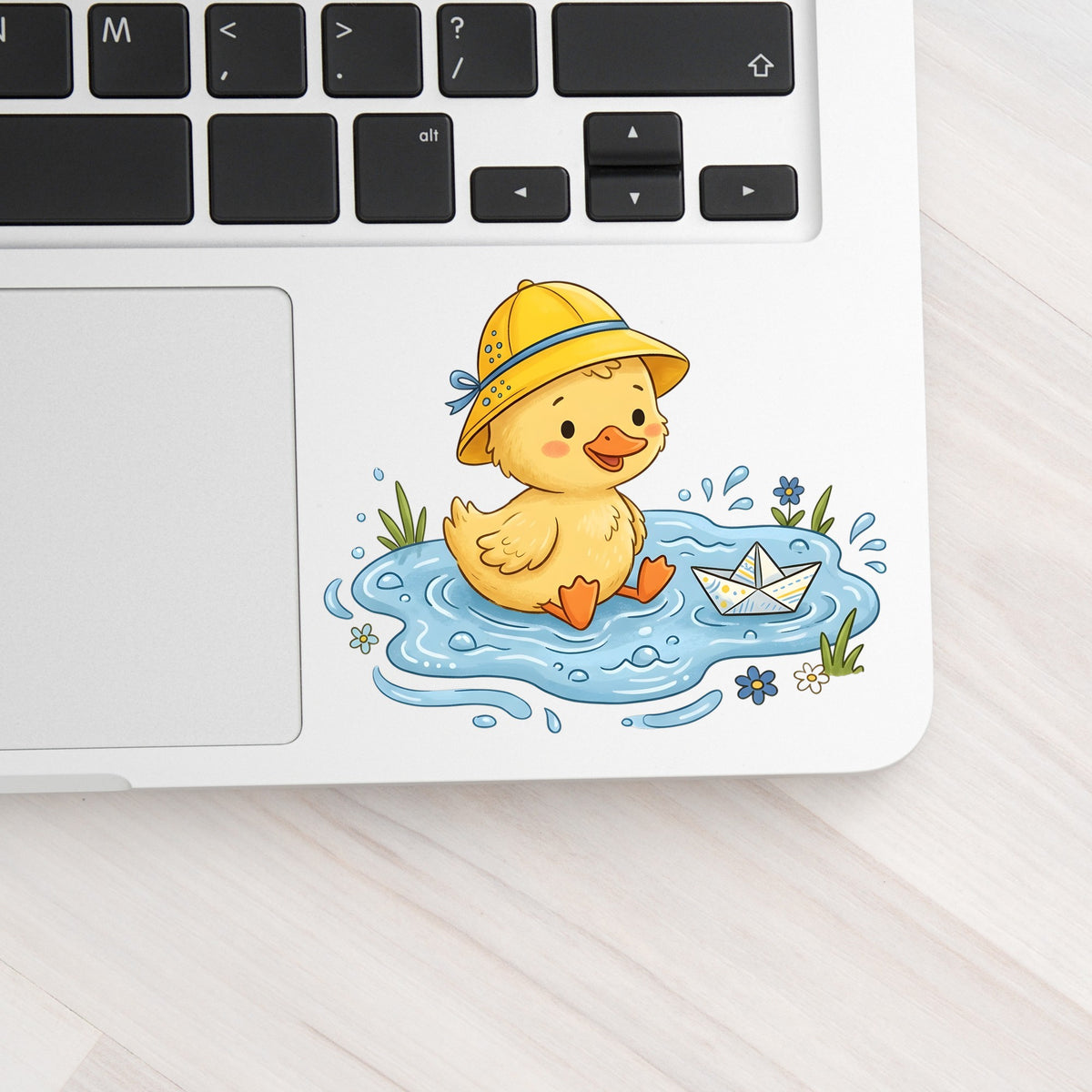Duckling In Rain Hat In Puddle UV DTF sticker applied to a silver laptop beside the keyboard
