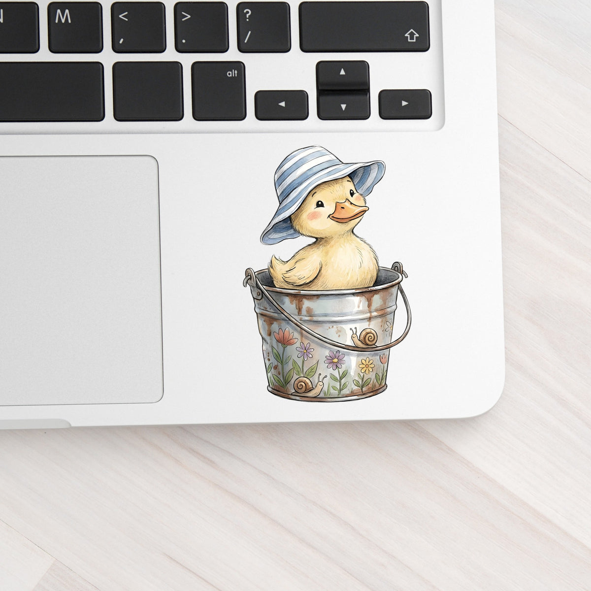 Duckling In Sun Hat In Bucket UV DTF sticker applied to a silver laptop beside the keyboard