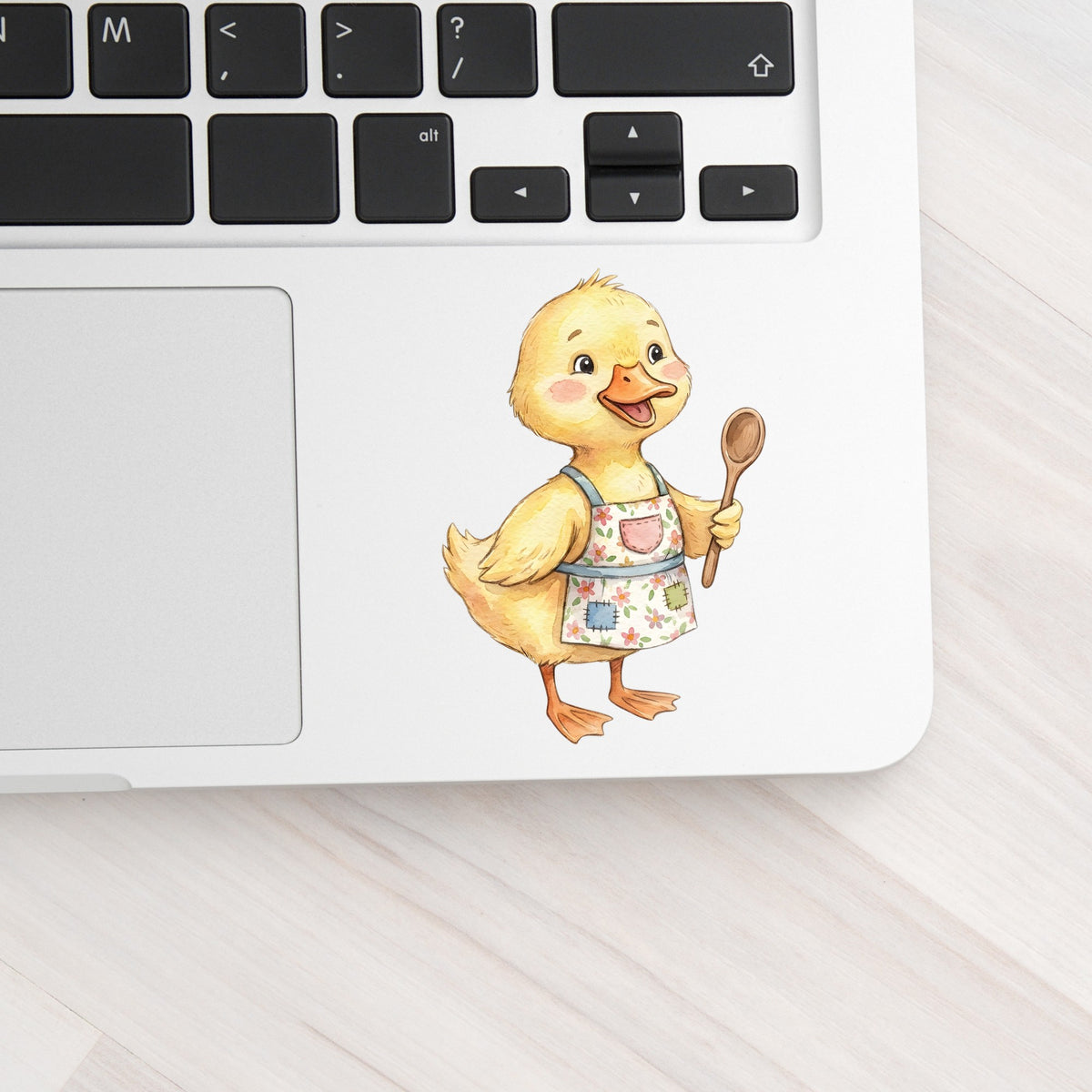 Duckling In Apron With Wooden Spoon UV DTF sticker applied to a silver laptop beside the keyboard