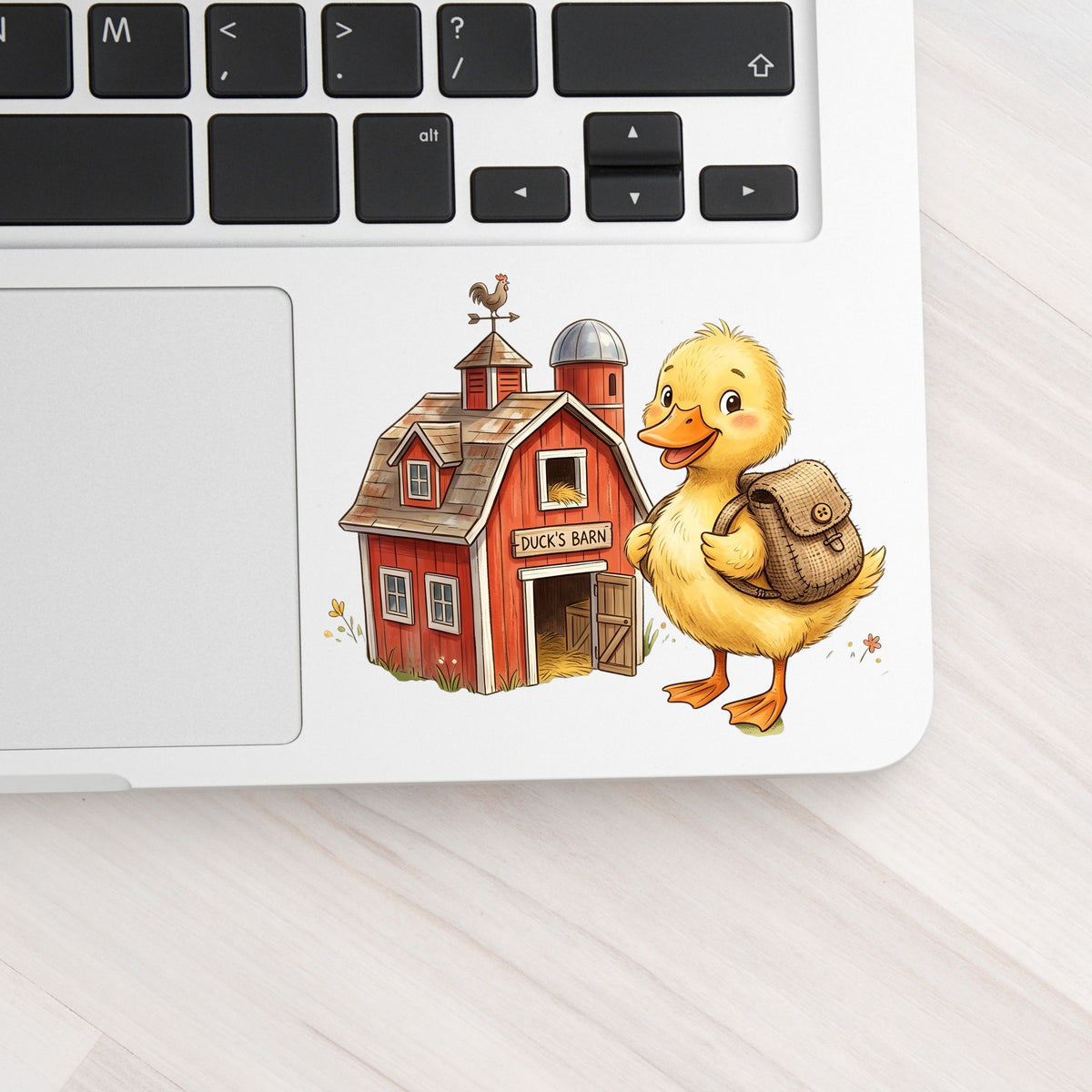 Duckling With Backpack And Red Barn UV DTF sticker applied to a silver laptop beside the keyboard