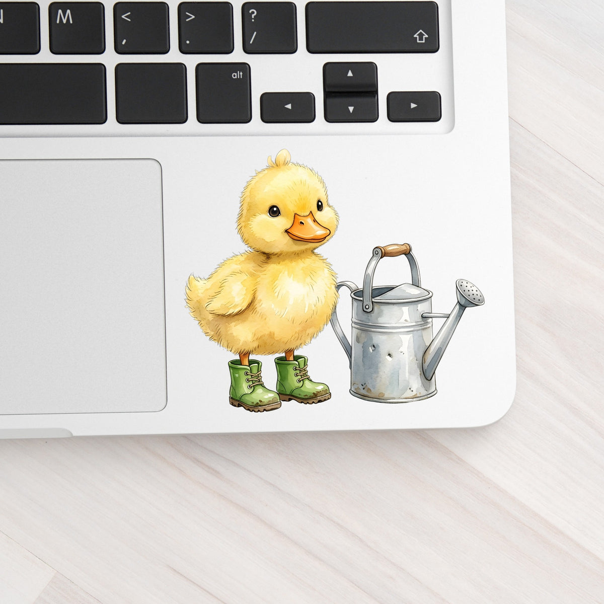 Duckling In Boots With Watering Can UV DTF sticker applied to a silver laptop beside the keyboard