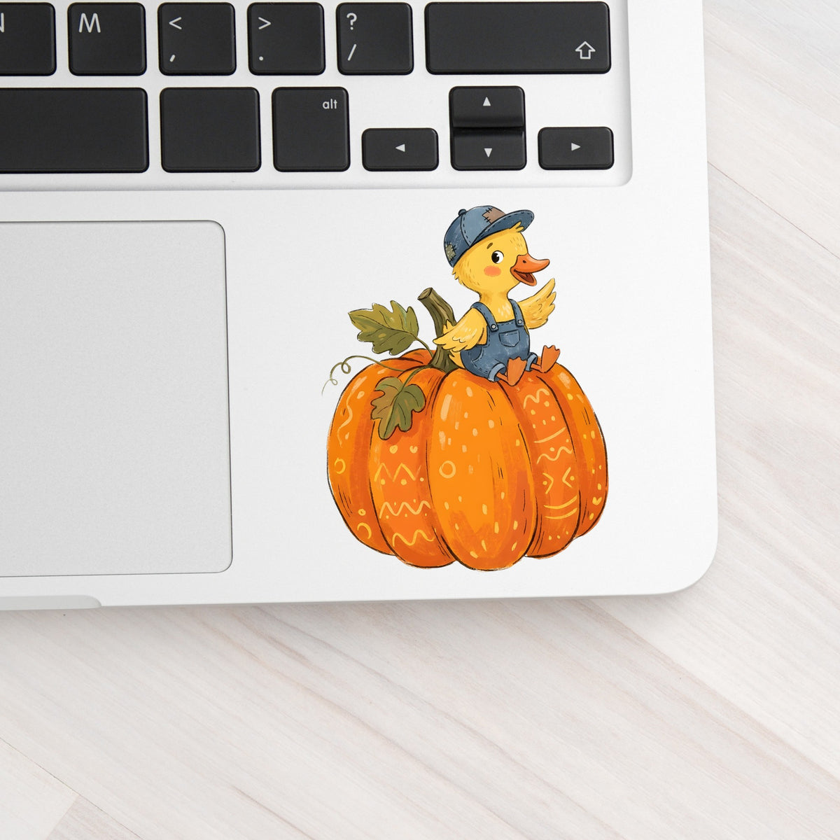 Duckling In Overalls On Pumpkin UV DTF sticker applied to a silver laptop beside the keyboard
