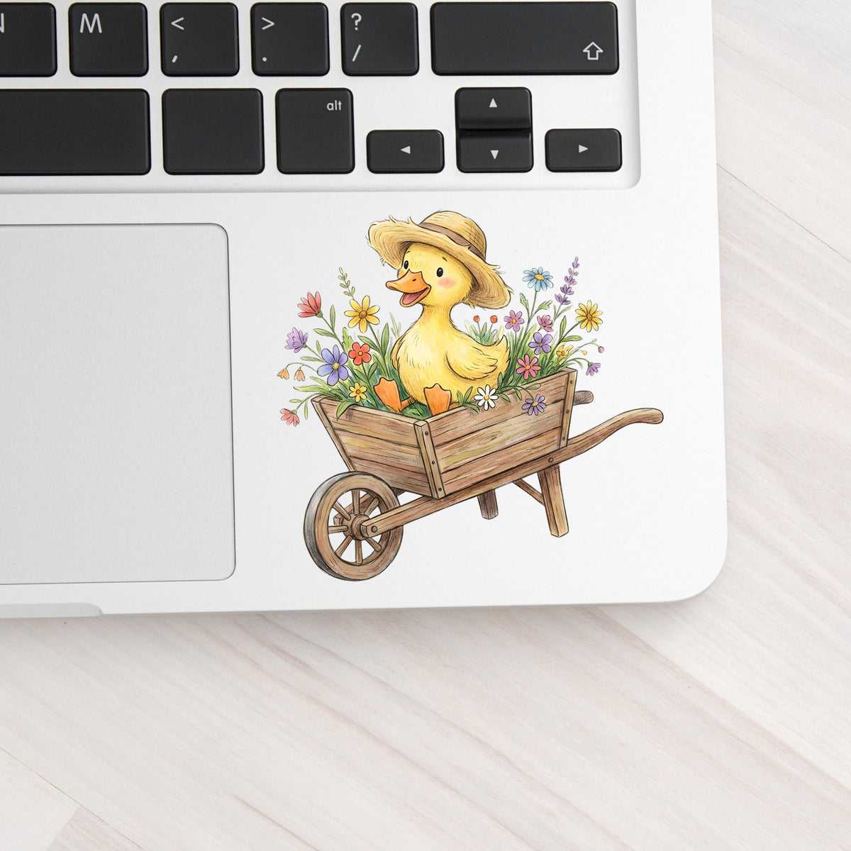 Duckling In Wheelbarrow Of Flowers UV DTF sticker applied to a silver laptop beside the keyboard