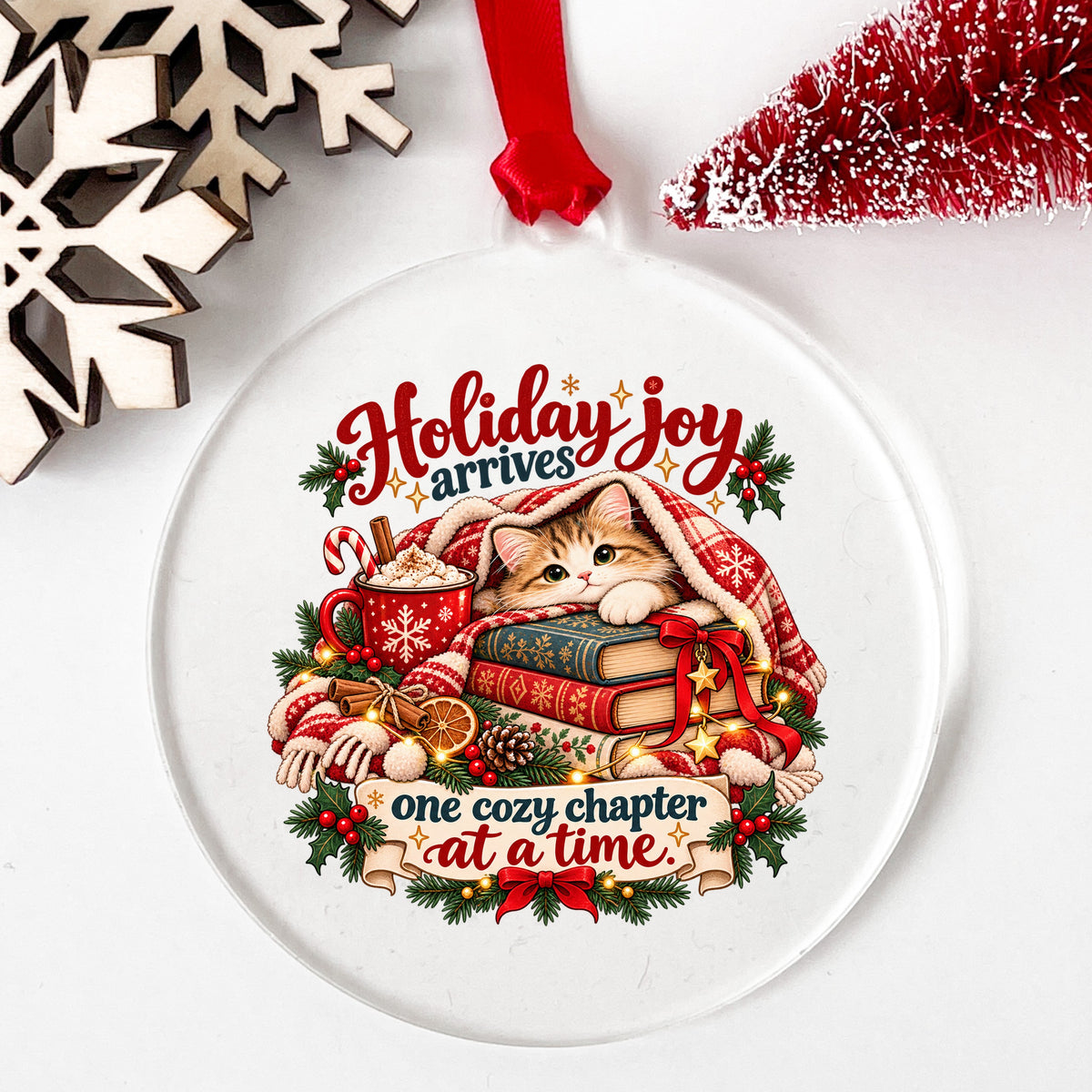 Cozy Kitten Under Plaid Blanket With Books UV DTF sticker applied to a clear round acrylic ornament with a red ribbon
