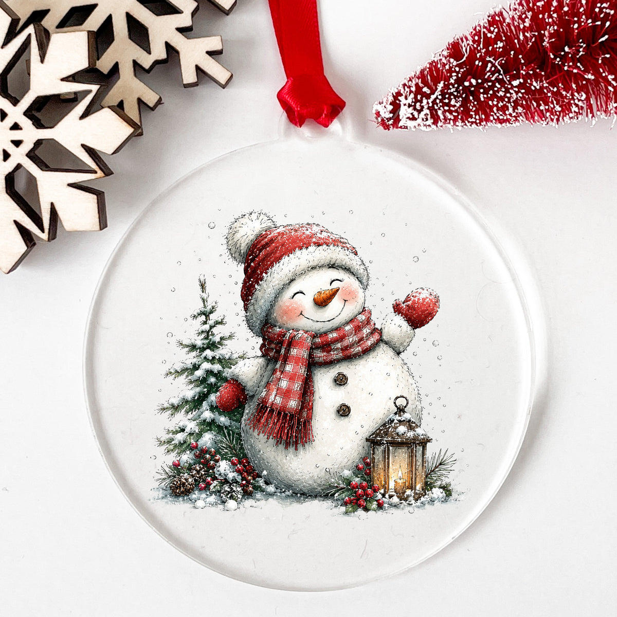 Waving Snowman By Snowy Pine Tree UV DTF sticker applied to a clear round acrylic ornament with a red ribbon