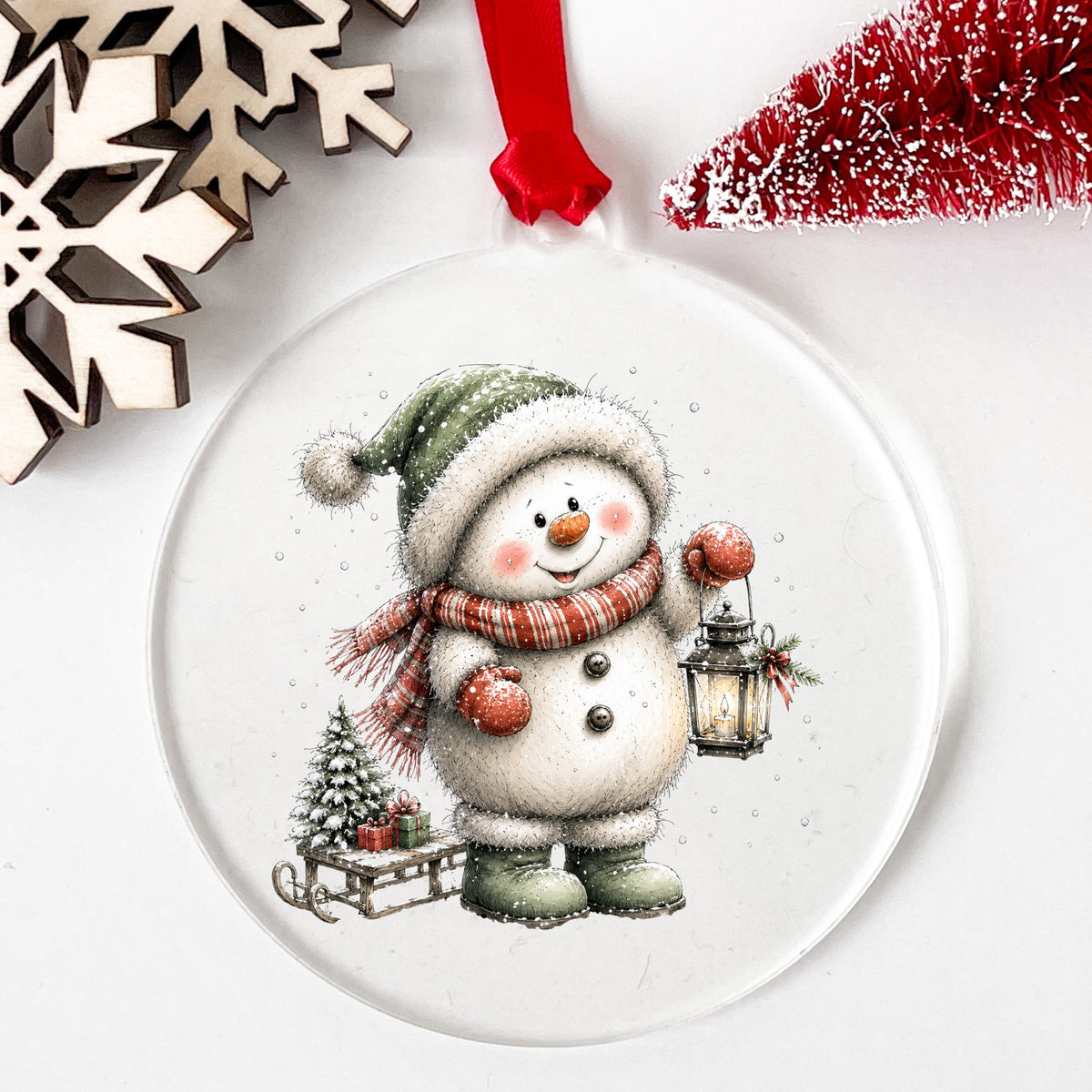 Green Hat Snowman With Sled And Gifts UV DTF sticker applied to a clear round acrylic ornament with a red ribbon