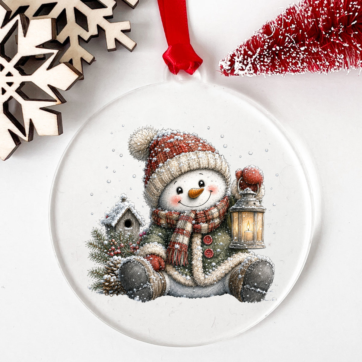 Sitting Snowman In Green Coat With Birdhouse UV DTF sticker applied to a clear round acrylic ornament with a red ribbon