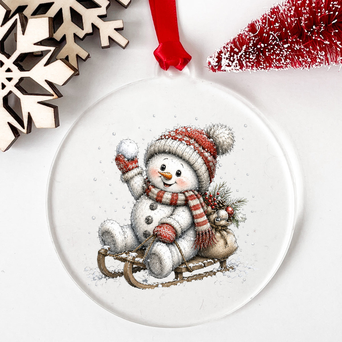 Snowball Snowman On Sled UV DTF sticker applied to a clear round acrylic ornament with a red ribbon