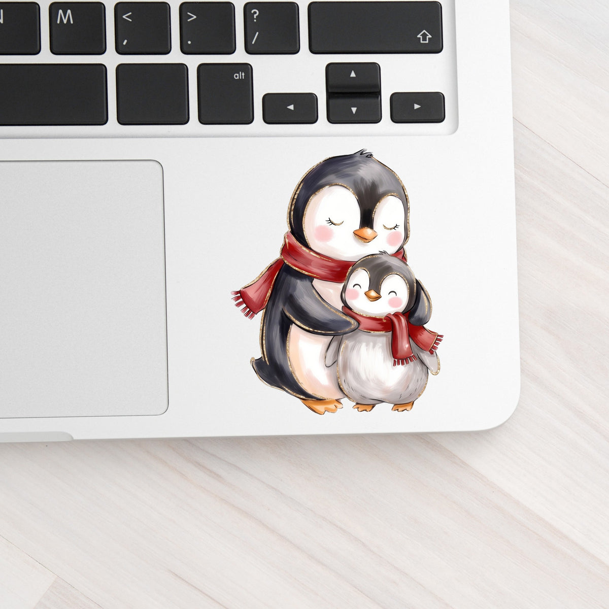 Mama And Baby Penguin In Red Scarves UV DTF sticker applied to a silver laptop beside the keyboard