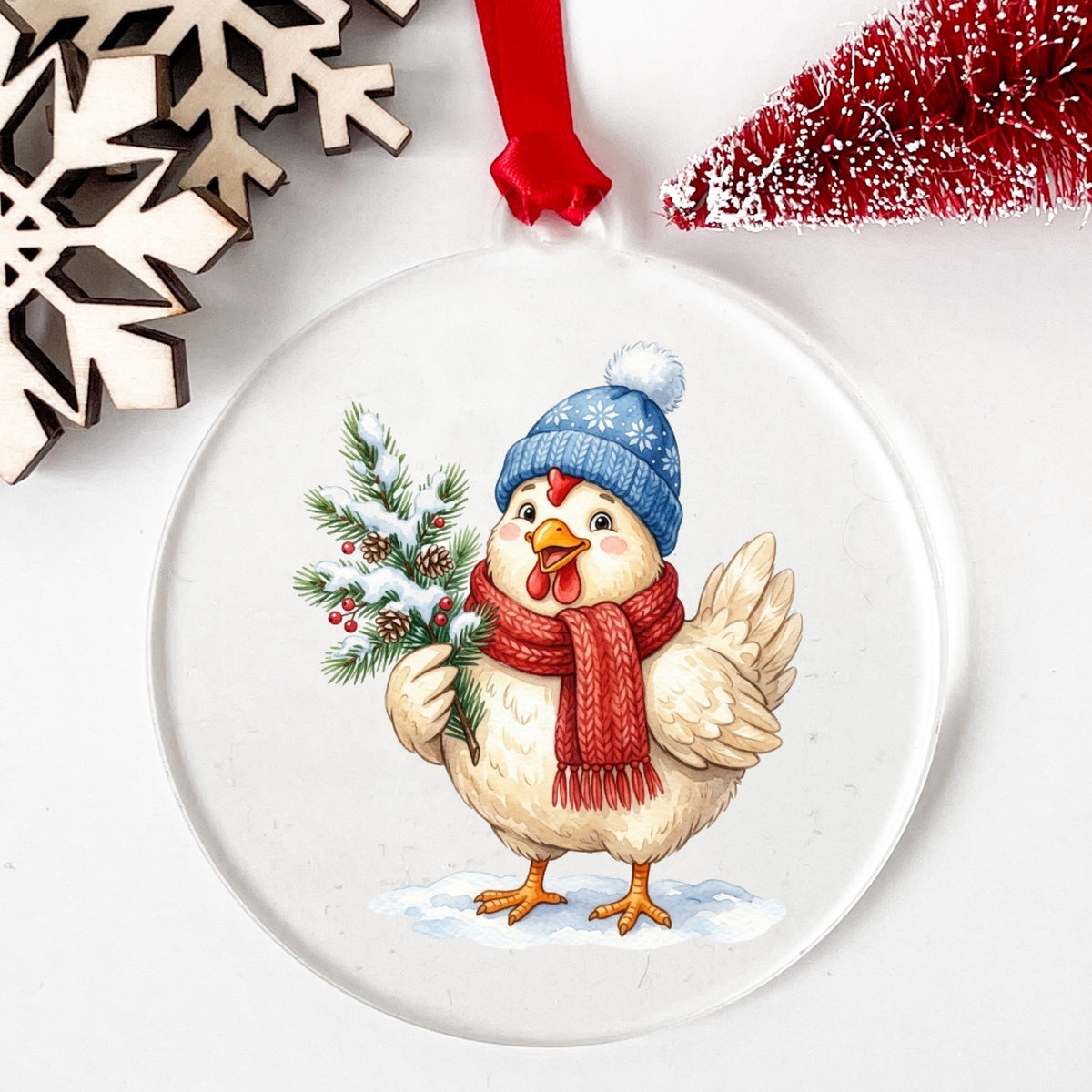 Blue Beanie Chicken With Pine Branch UV DTF sticker applied to a clear round acrylic ornament with a red ribbon