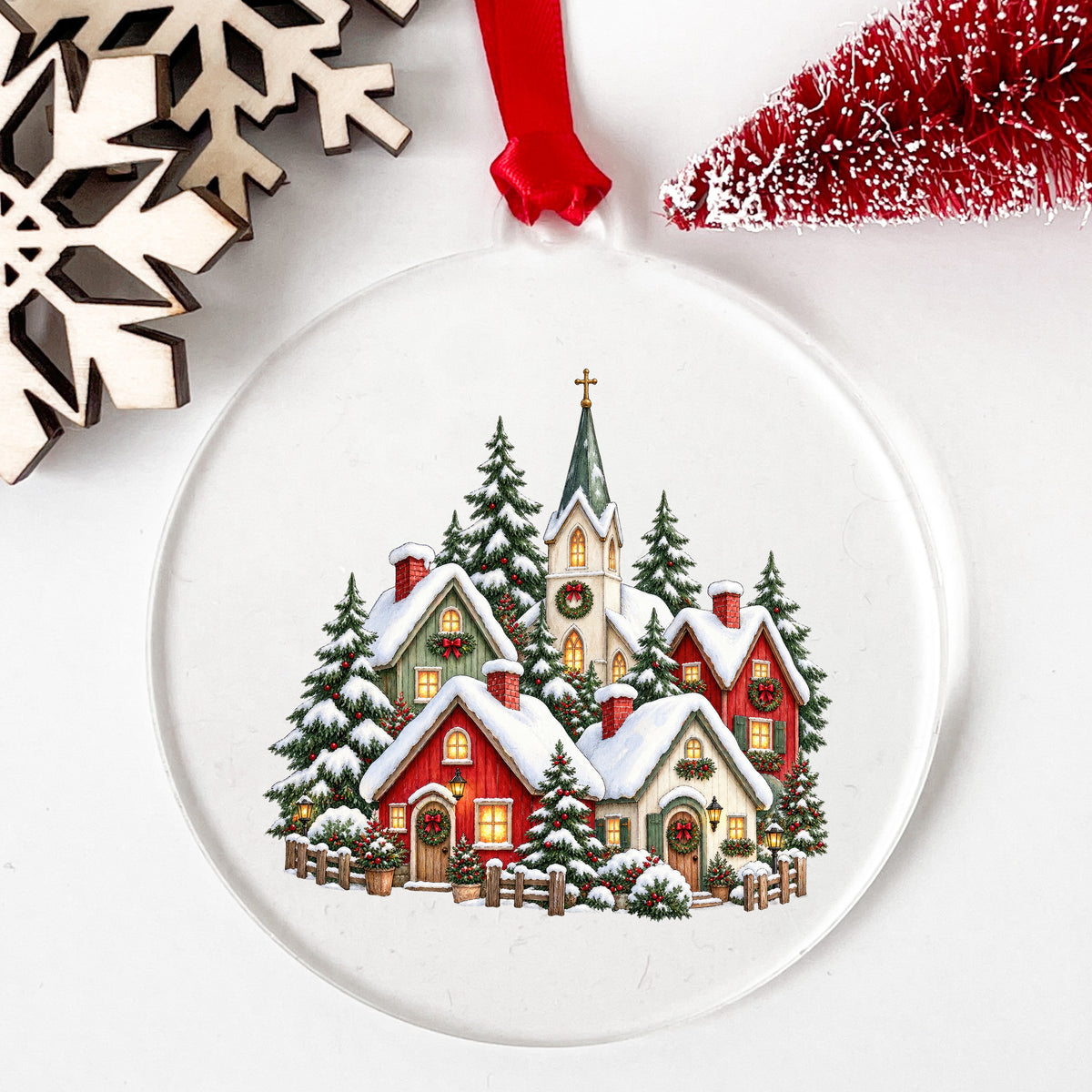 Snowy Christmas Village With Church UV DTF sticker applied to a clear round acrylic ornament with a red ribbon
