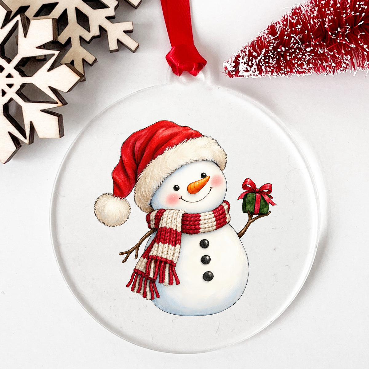 Snowman In Santa Hat With Gift UV DTF sticker applied to a clear round acrylic ornament with a red ribbon