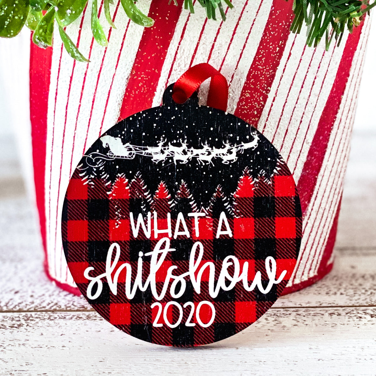 What a shitshow on round ornament. Red and black buffalo plaid background with Santa&#39;s sleigh